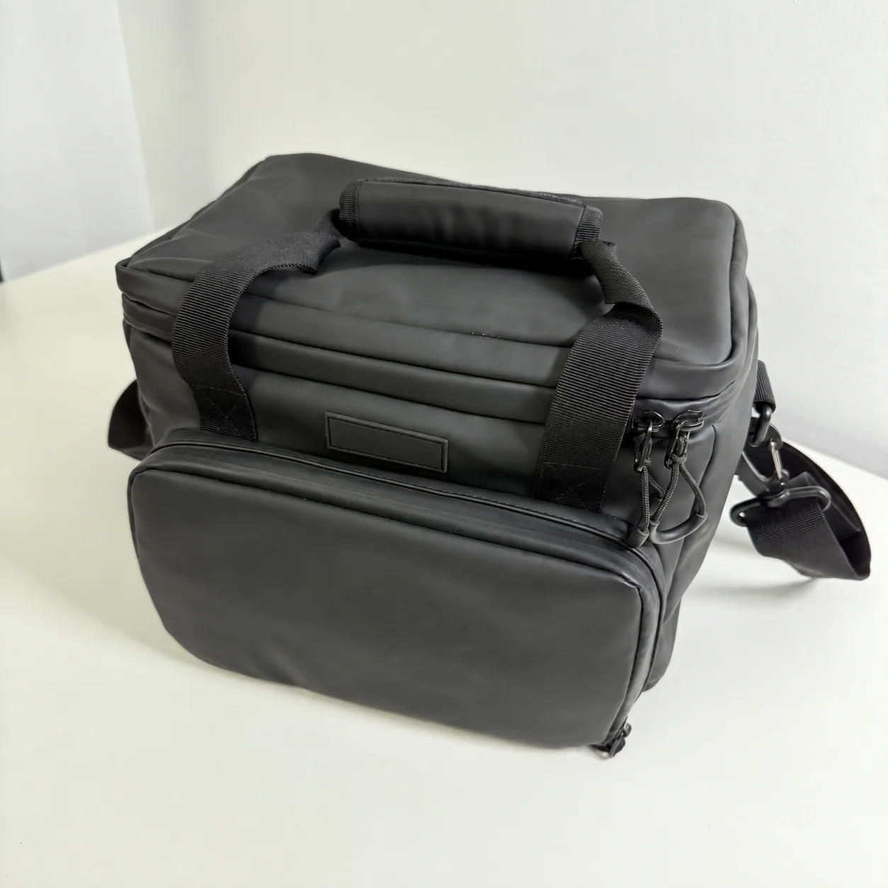 Three-quarter view of the actual black soft cooler sample showing its padded top handle, zippered front pocket, straps and zipper pulls; customer logo removed